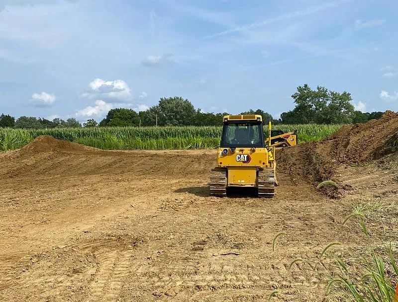 Yellow bulldozer on dirt, clearing land near a field of tall green plants under a blue sky.