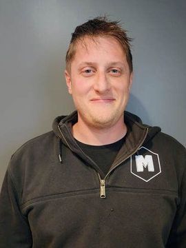 Man with short brown hair, smiling, wearing a black hooded sweatshirt with a white logo in front of a gray wall.