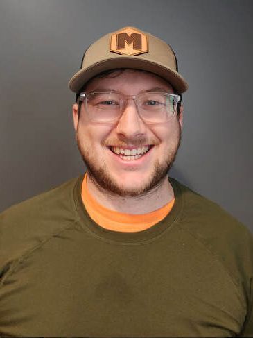Man wearing glasses, hat, and green shirt smiling in front of a gray backdrop.