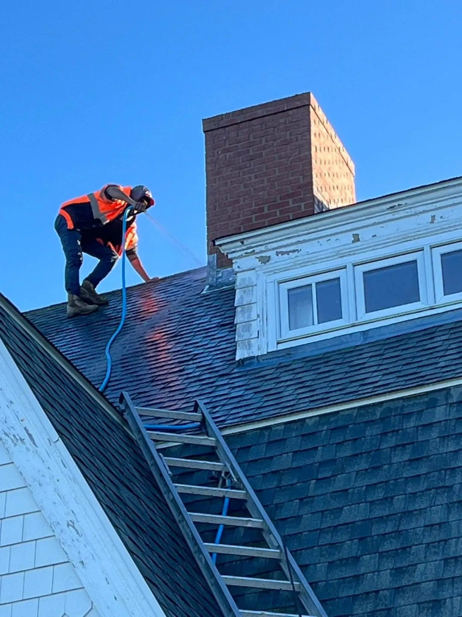 Worker on a steep roof securing a ladder beside a brick chimney and white dormer windows.