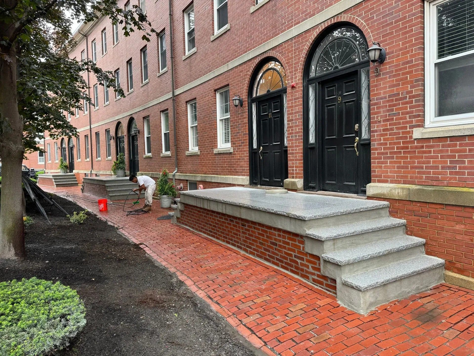 Brick townhouse entrance with black doors, concrete steps, and a red brick walkway on a rainy day.