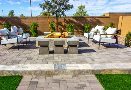Modern patio with white sofas, gray chairs, and a fire pit on paver stones in a landscaped backyard