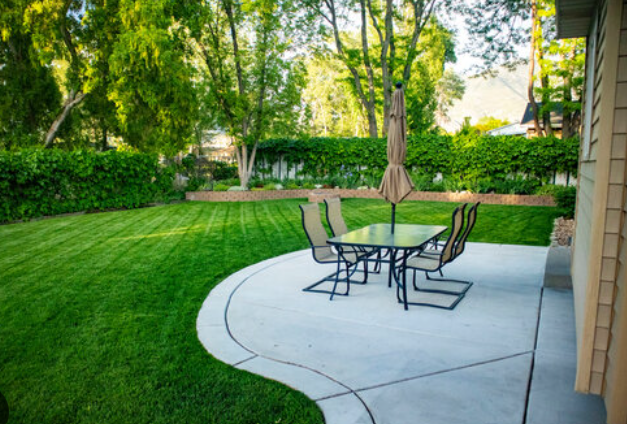 Patio table and chairs on a curved concrete pad beside a green lawn and trees