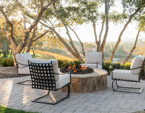 Outdoor patio with four lounge chairs around a stone fire pit at sunset, surrounded by trees