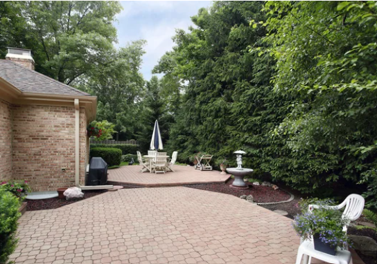 Brick patio with outdoor furniture, white umbrella, and lush green trees along the yard edge