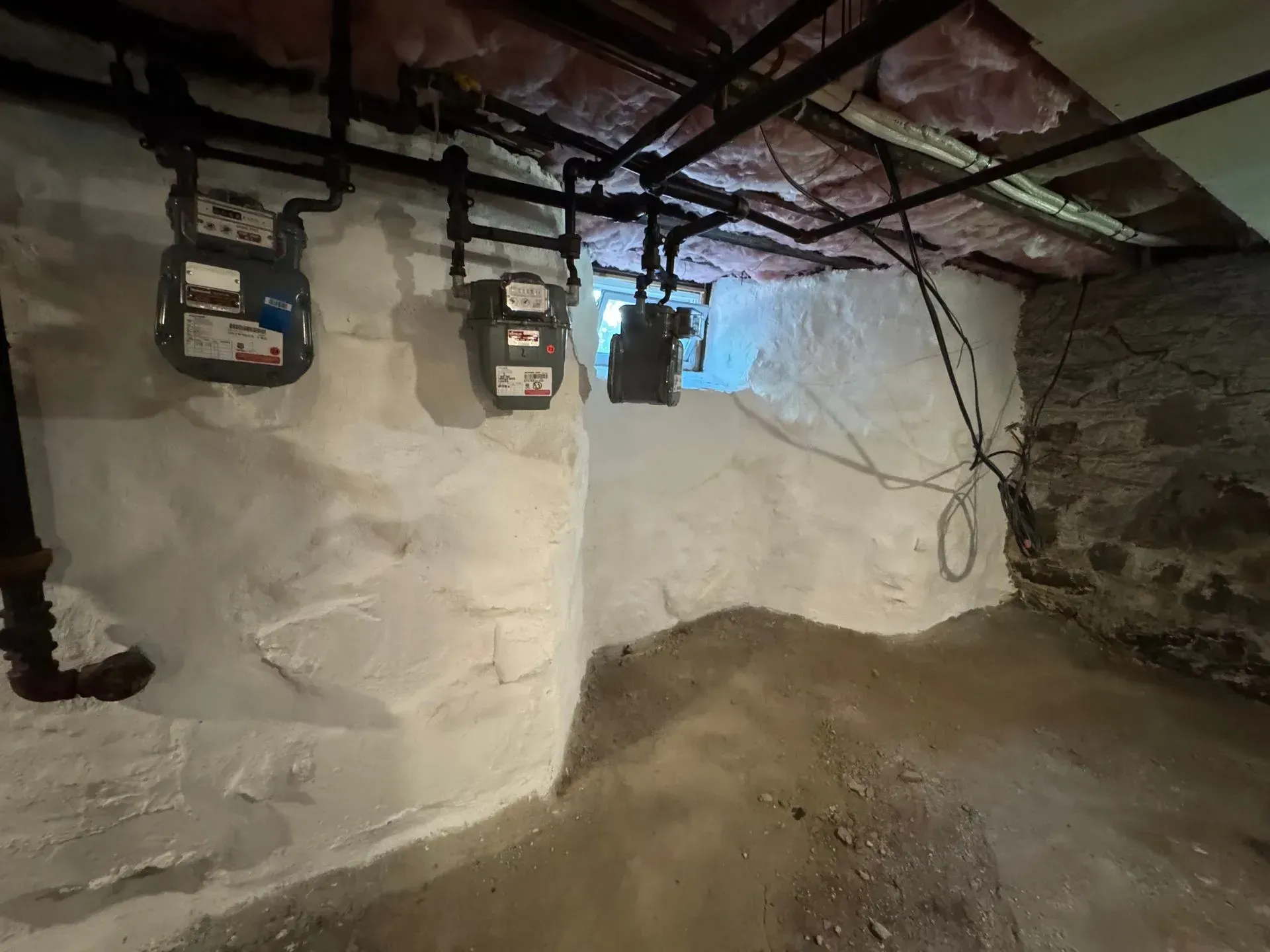Basement interior with three gas meters on white-painted stone wall. Black pipes, unfinished ceiling.