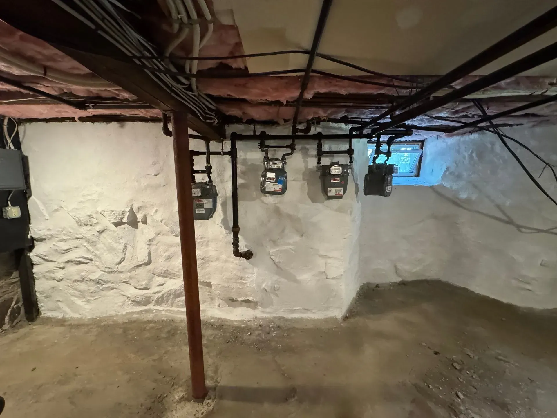 Basement with gas meters on a white stone wall, exposed pipes and beams overhead.