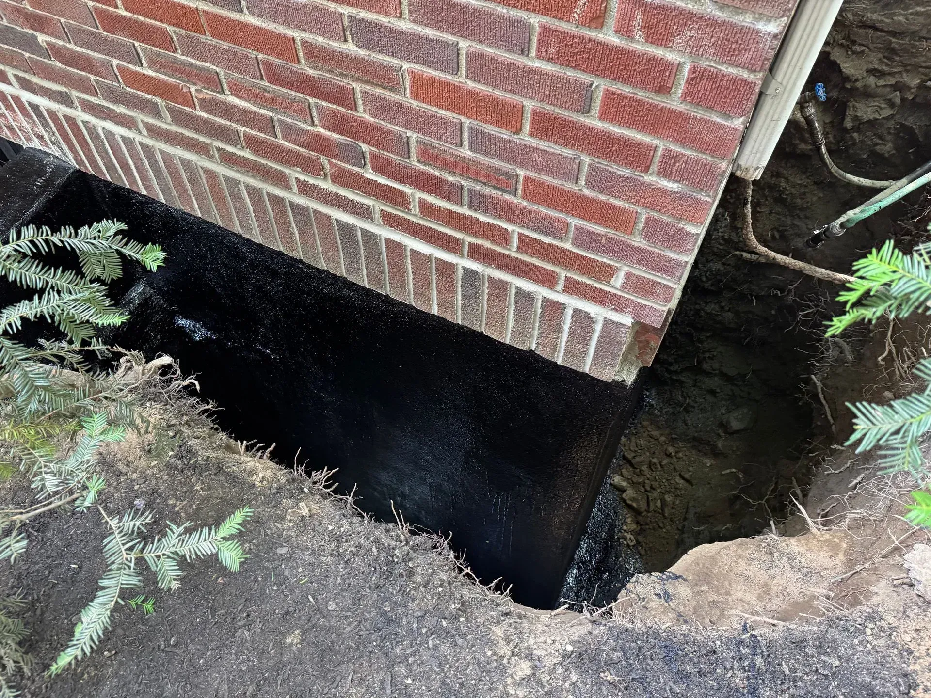 Foundation trench next to a brick wall coated with a black waterproofing material.