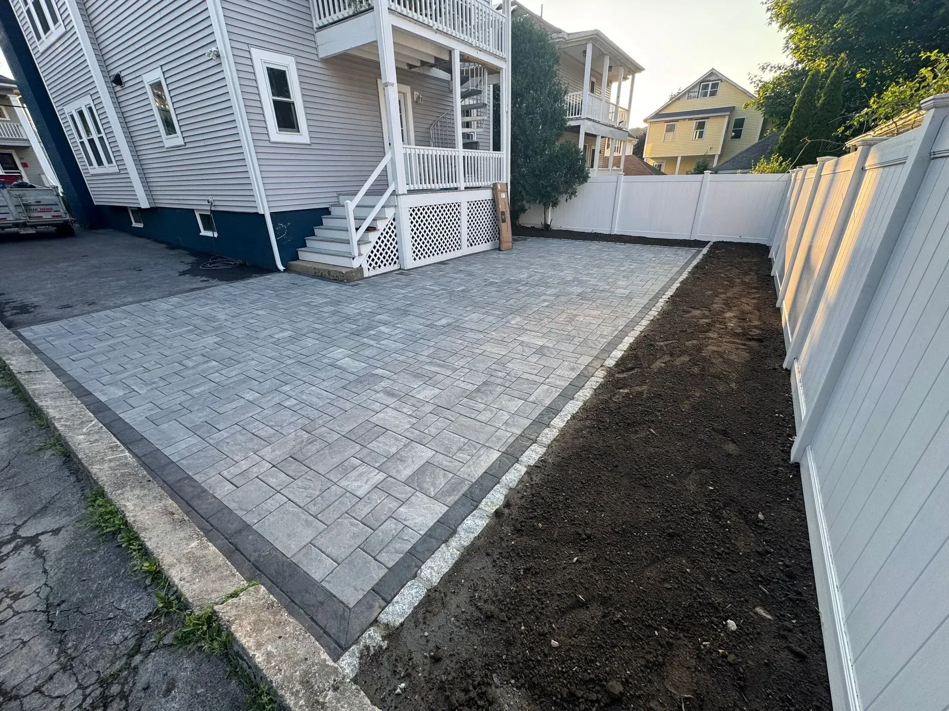 Gray paver driveway beside a fenced yard, next to a multi-story house.