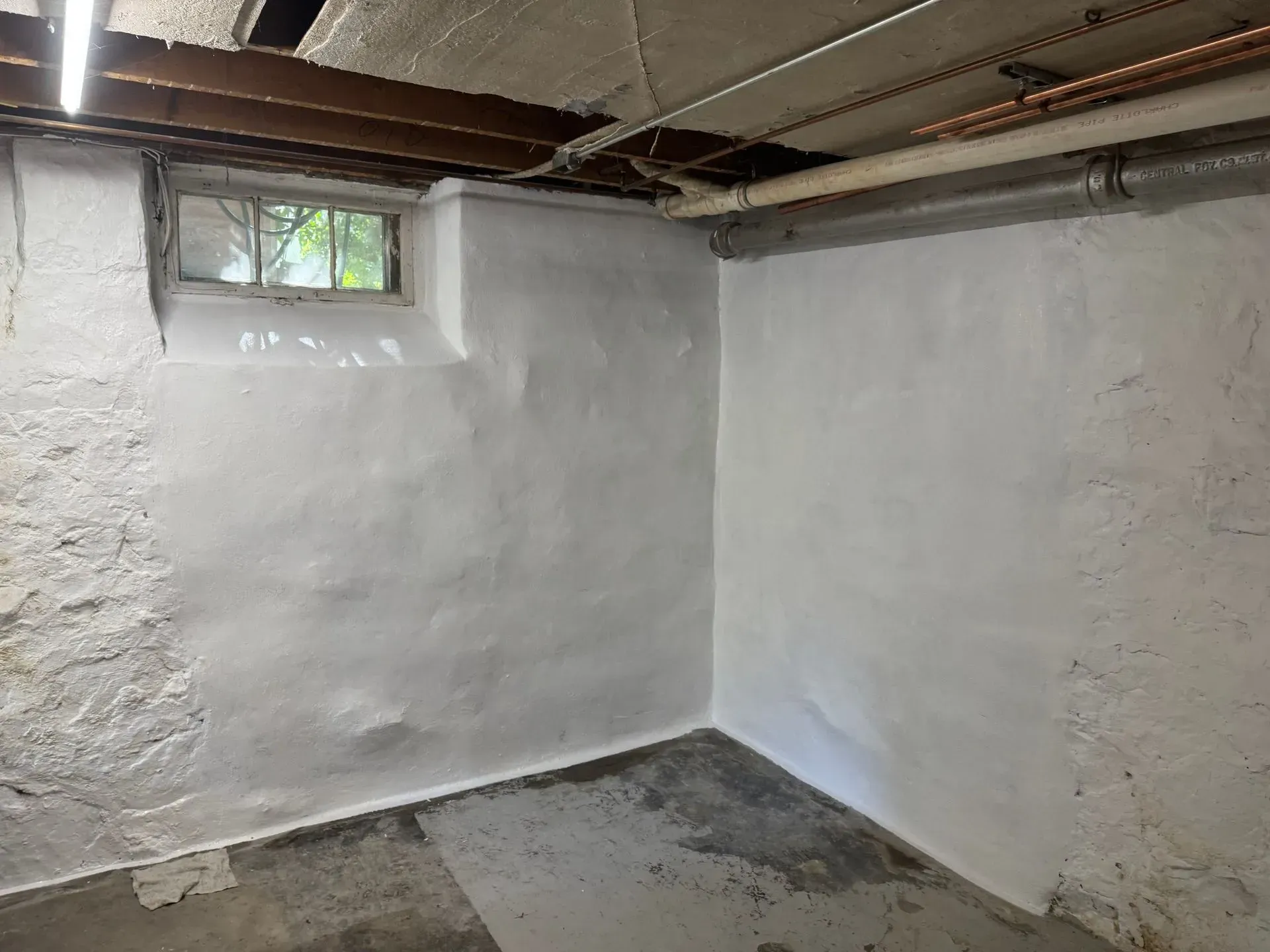 Basement interior, white-painted concrete walls, small window, visible pipes and ductwork, concrete floor.