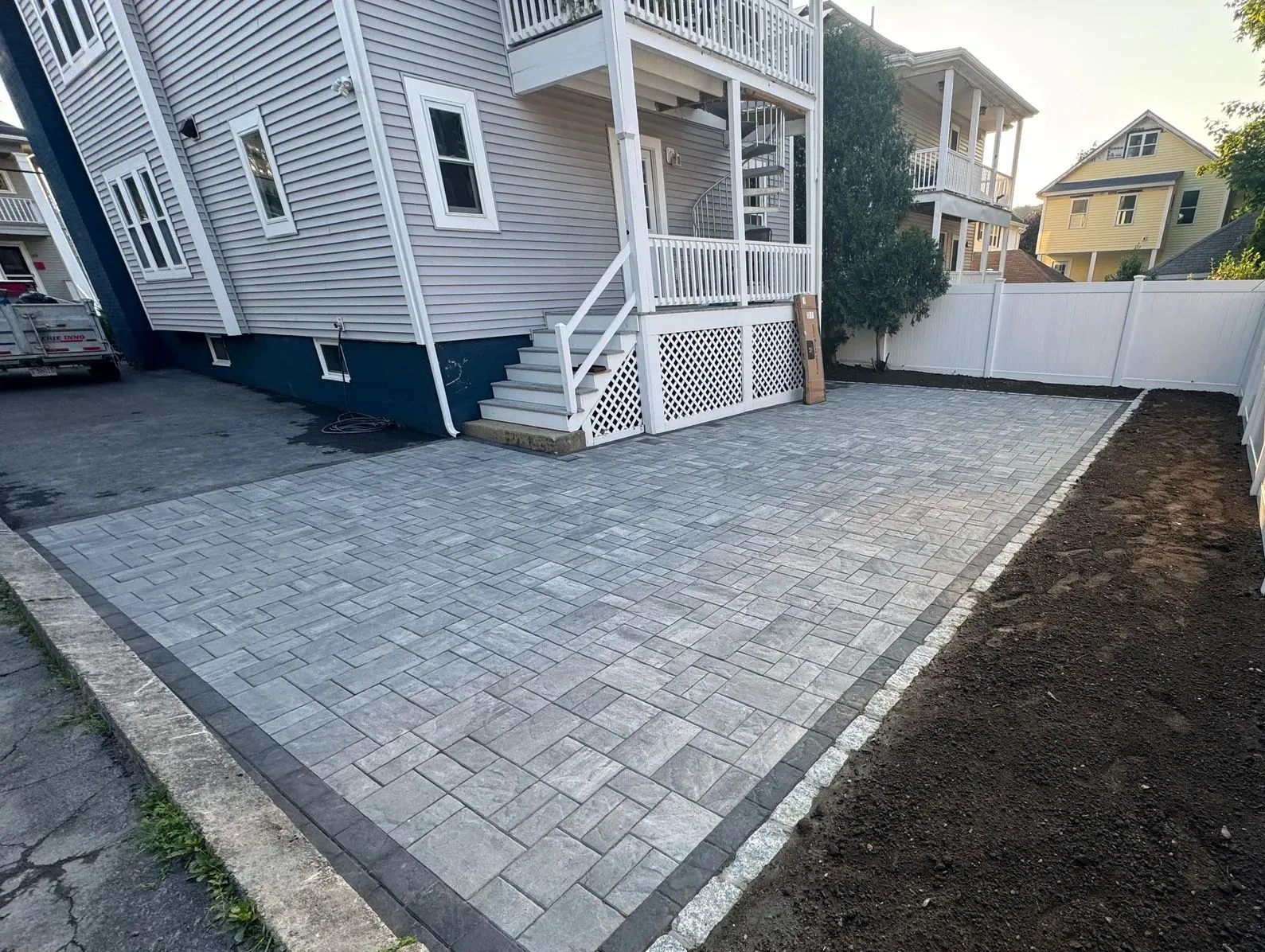 Paved backyard patio beside a gray house, bordered by a white fence and bare soil strip