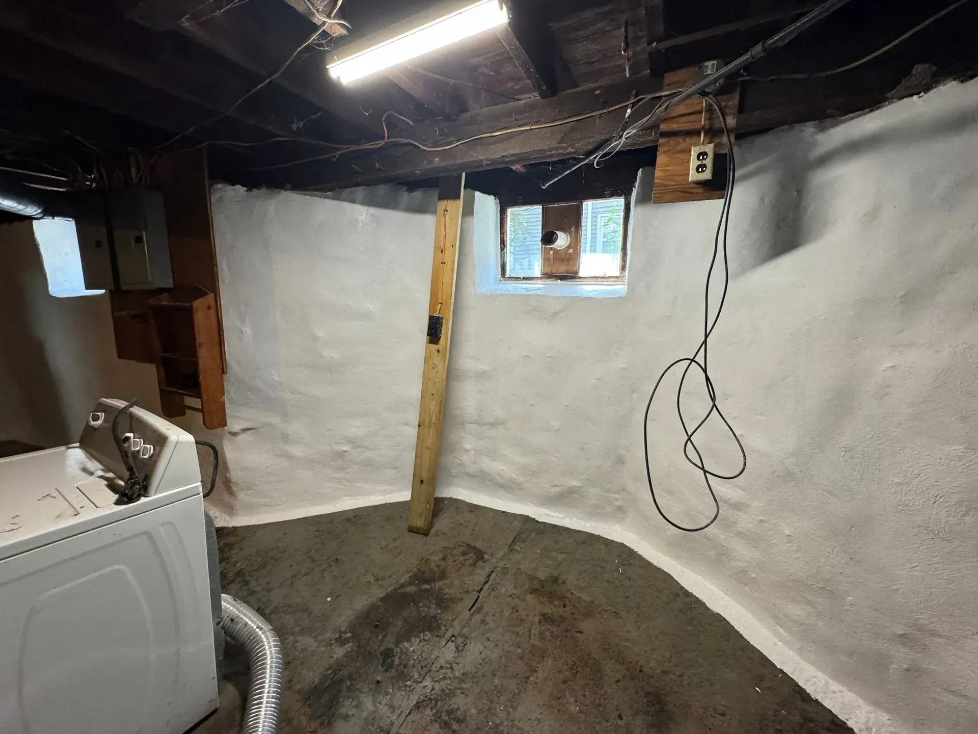 Basement interior with white walls, unfinished ceiling, washing machine. A window with a wooden beam prop.