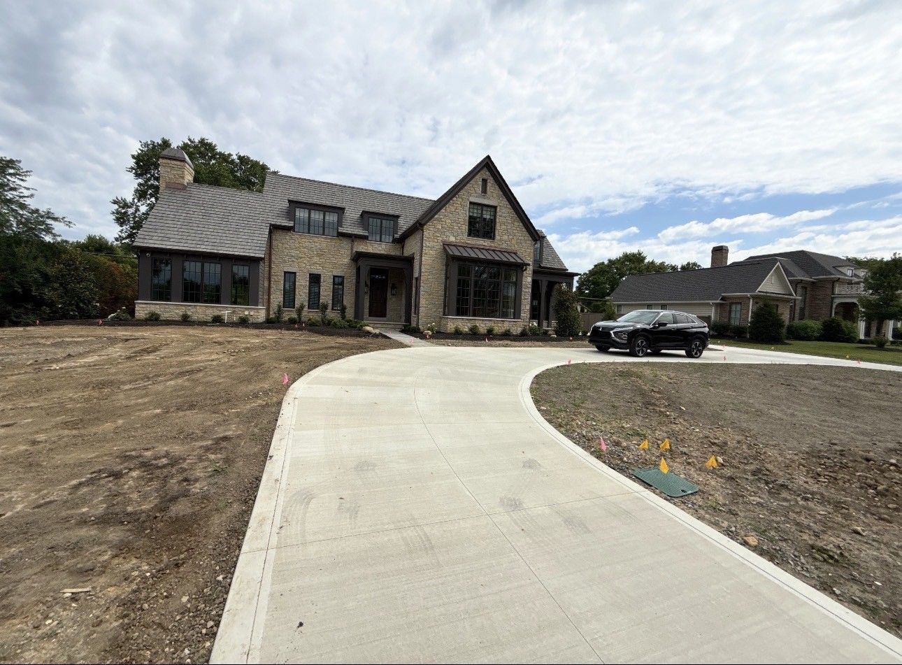 Stone house with steep roof, curved driveway, and parked SUV under a cloudy sky