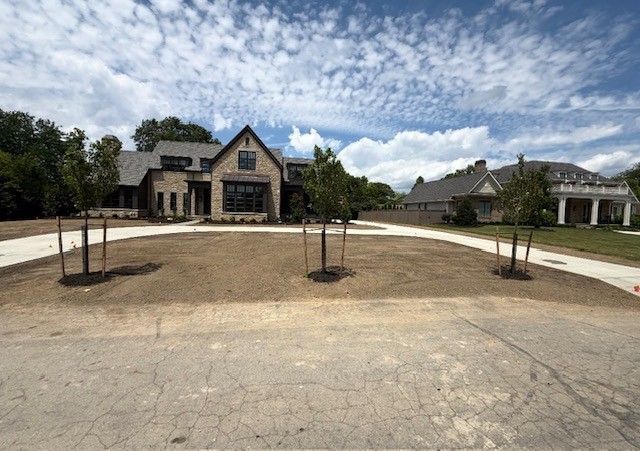 Modern stone house with a wide front drive, young trees, and a partly cloudy sky