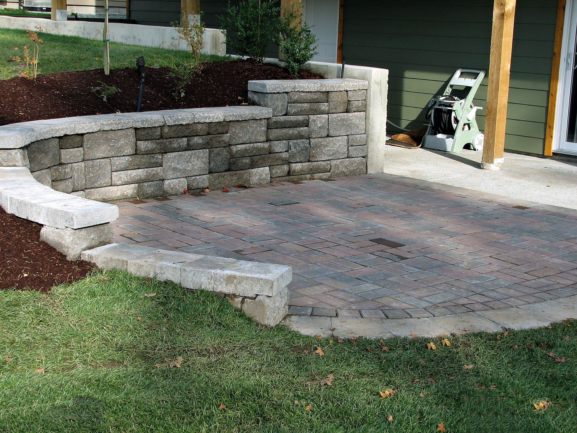 Stone patio with curved retaining wall beside a grassy yard and house garage.