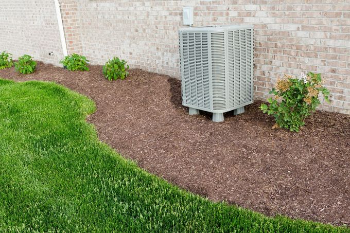 Air conditioning unit beside a brick wall, with mulch bed and green lawn in front