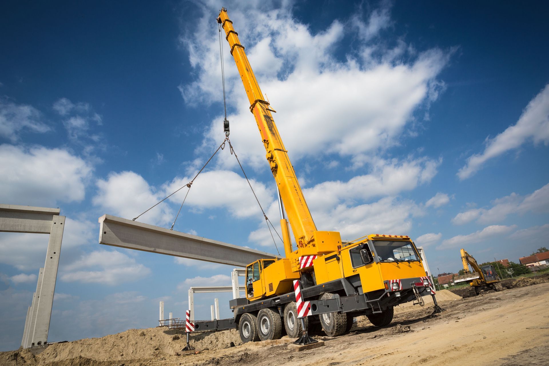 Yellow crane lifting a long concrete beam at a construction site against a cloudy blue sky.