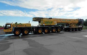 Yellow mobile crane and trailer on a paved surface under a blue sky with clouds.