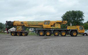 Yellow crane on a trailer, parked on gravel. A second truck is parked next to it. Overcast sky.