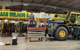 Two people on a lift working on a large yellow crane in a warehouse; the crane says "A&B Welding."