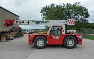 Red crane truck with its arm extended, parked in front of a building with a person inside.