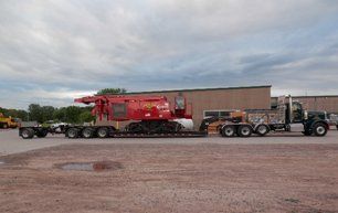 Red industrial machine on a flatbed trailer being pulled by a semi-truck on a paved lot.