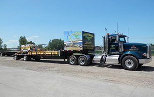 Blue semi-truck pulling a flatbed trailer loaded with equipment, parked on gravel in front of a blue sky.
