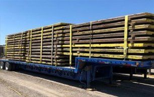 Wooden planks stacked and secured on a blue flatbed trailer, yellow straps, outdoors.