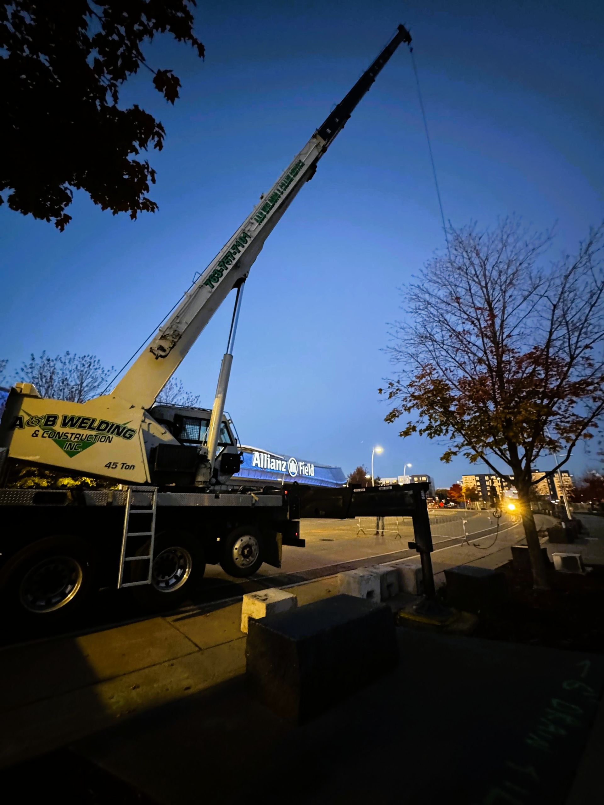 A crane lifting something at dusk. The crane is on a truck, with a tree and streetlights in the background.