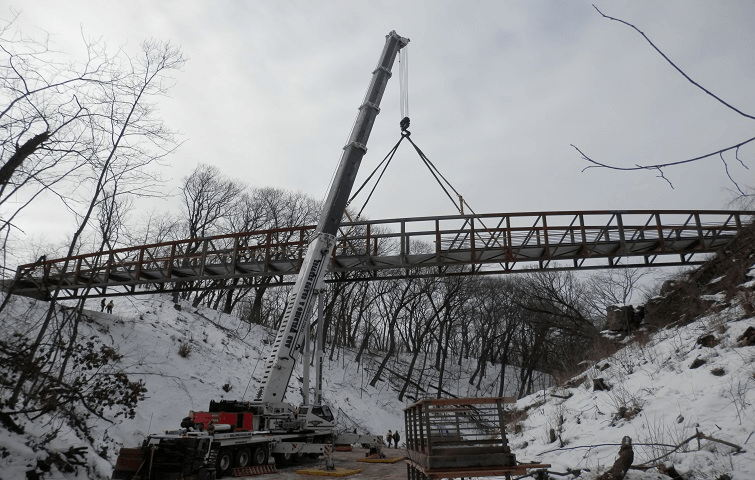 Crane lifting a long metal bridge section over a snow-covered ravine in a wooded area.