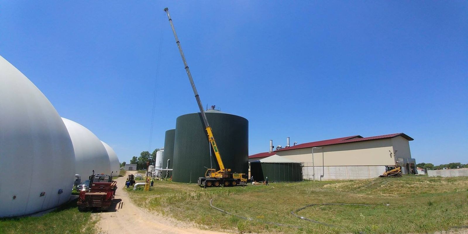 Construction site with a crane lifting something onto a large, dark green cylindrical tank. Blue sky.