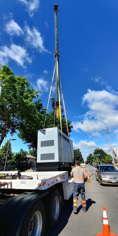 A large generator is being lifted from a flatbed truck by a crane on a sunny street.