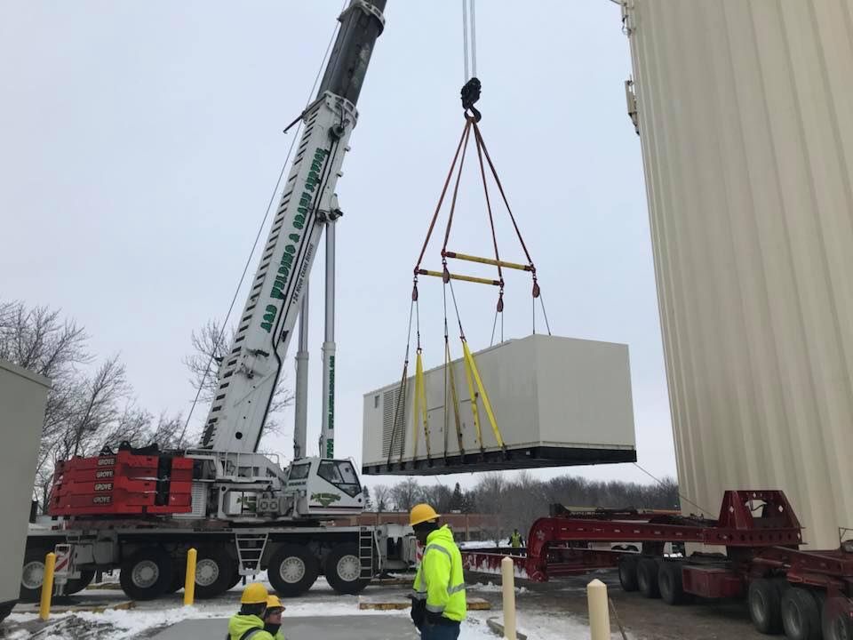 A crane lifting a large, rectangular beige unit at a construction site; workers in safety vests nearby.