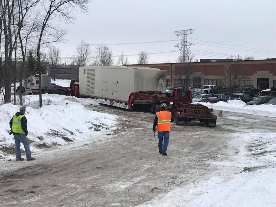 Workers guide a large, rectangular generator on a flatbed trailer in a snowy industrial area.