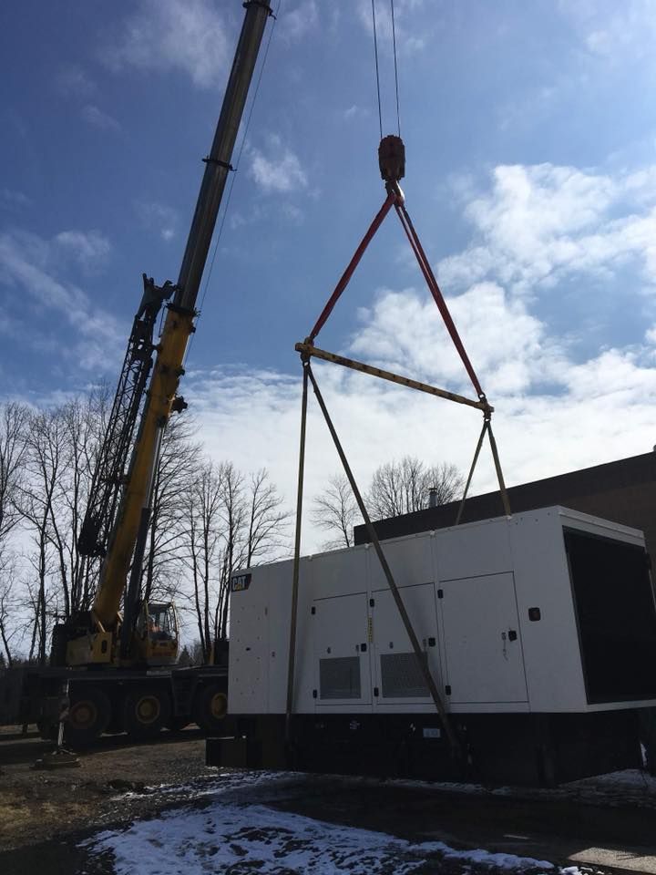 A crane lifting a large white generator. Sunny day, snow on the ground, trees in the background.