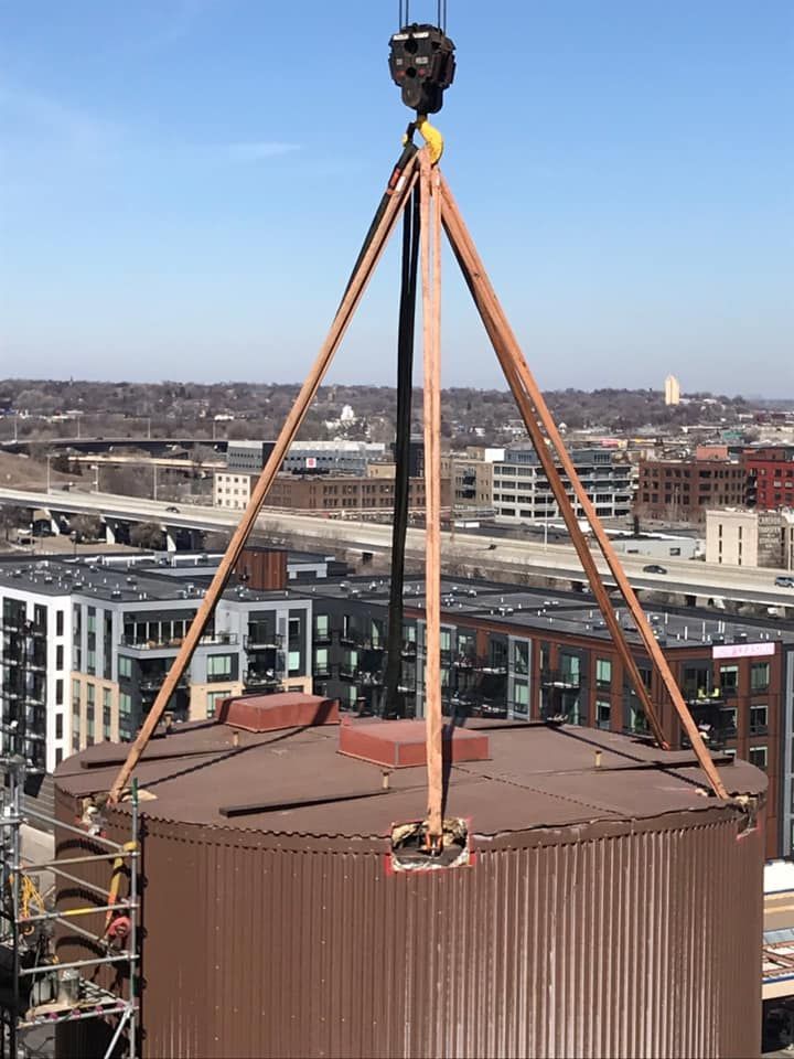 Crane lifting a large, brown cylindrical structure with a rooftop, possibly a water tank, against a city backdrop.