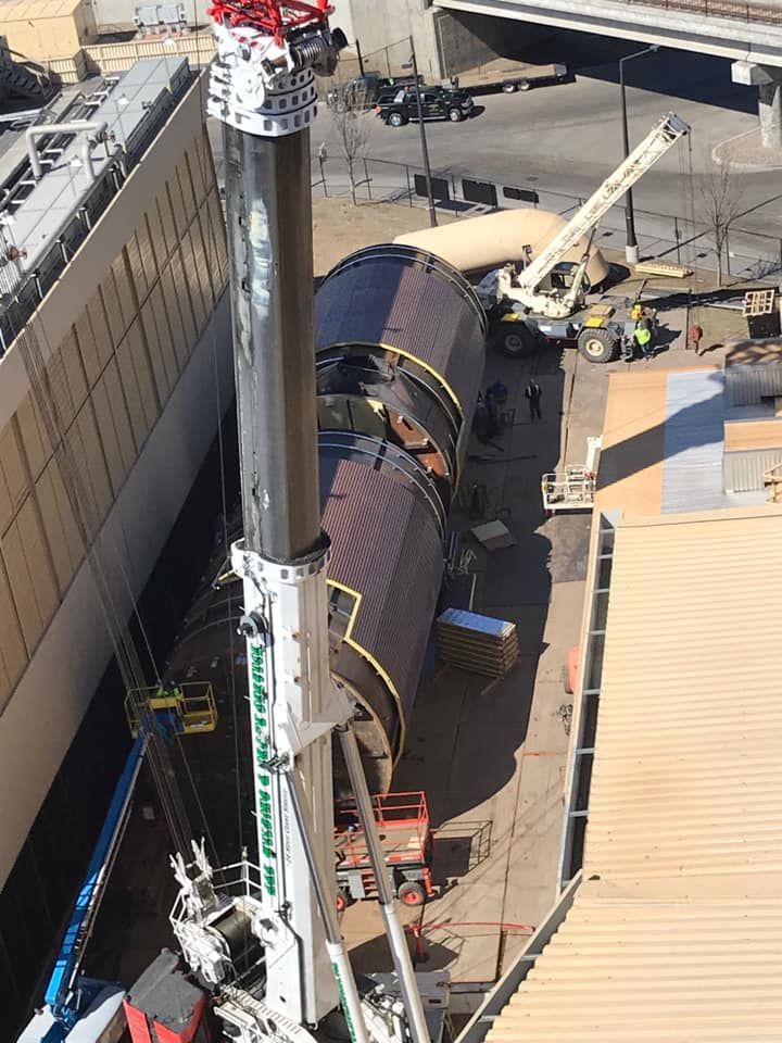 Crane lifting large cylindrical structure next to a building, possibly construction site.