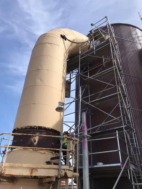 Scaffolding next to a large tan pipe and reddish-brown industrial silo; worker on a platform.