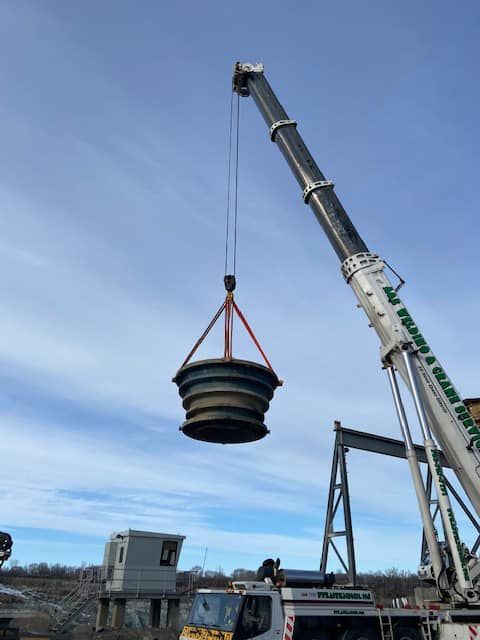 A crane lifting a large, dark-colored, cylindrical object against a cloudy sky.