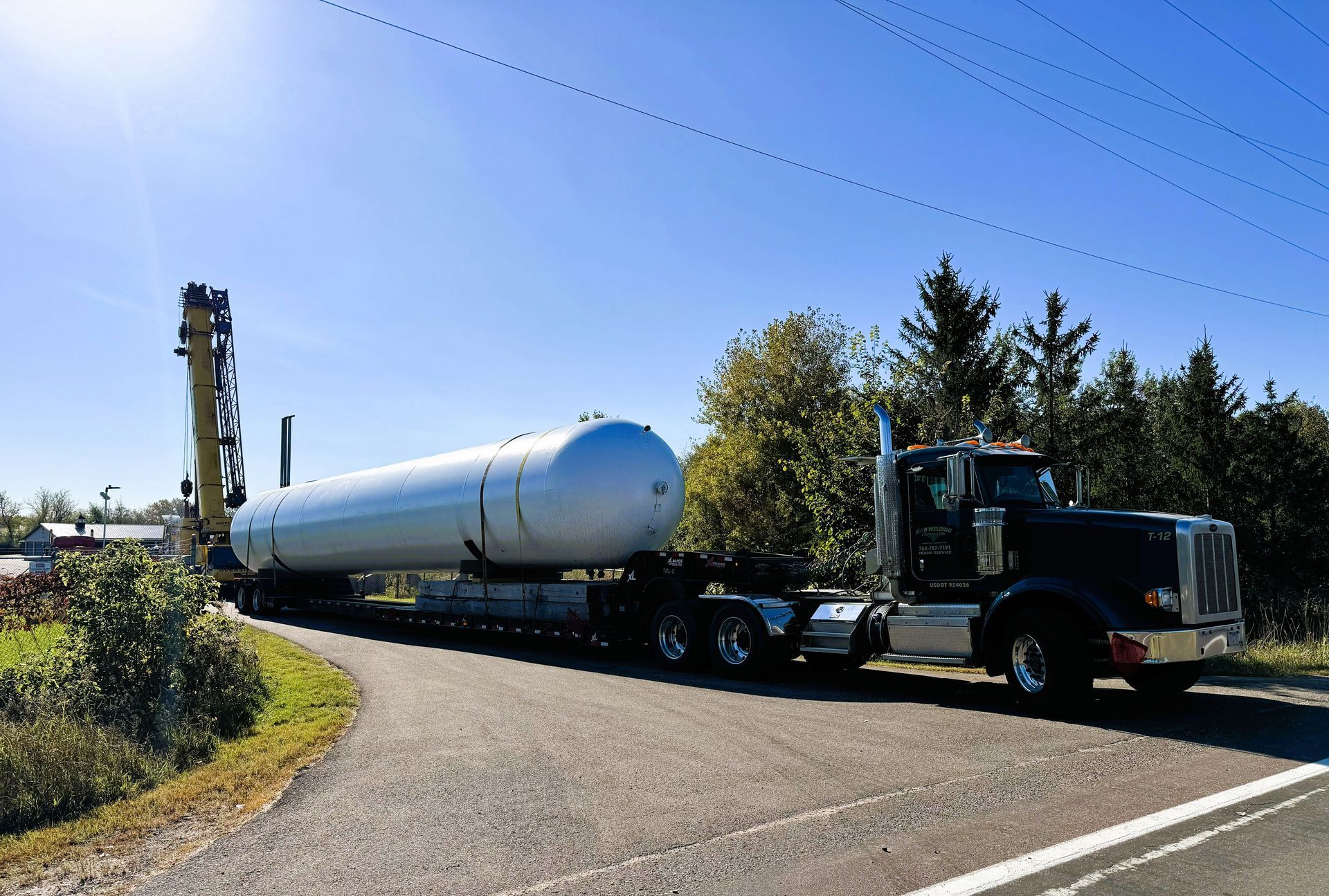 Semi-truck hauling a large white tank along a road, trees and blue sky visible.