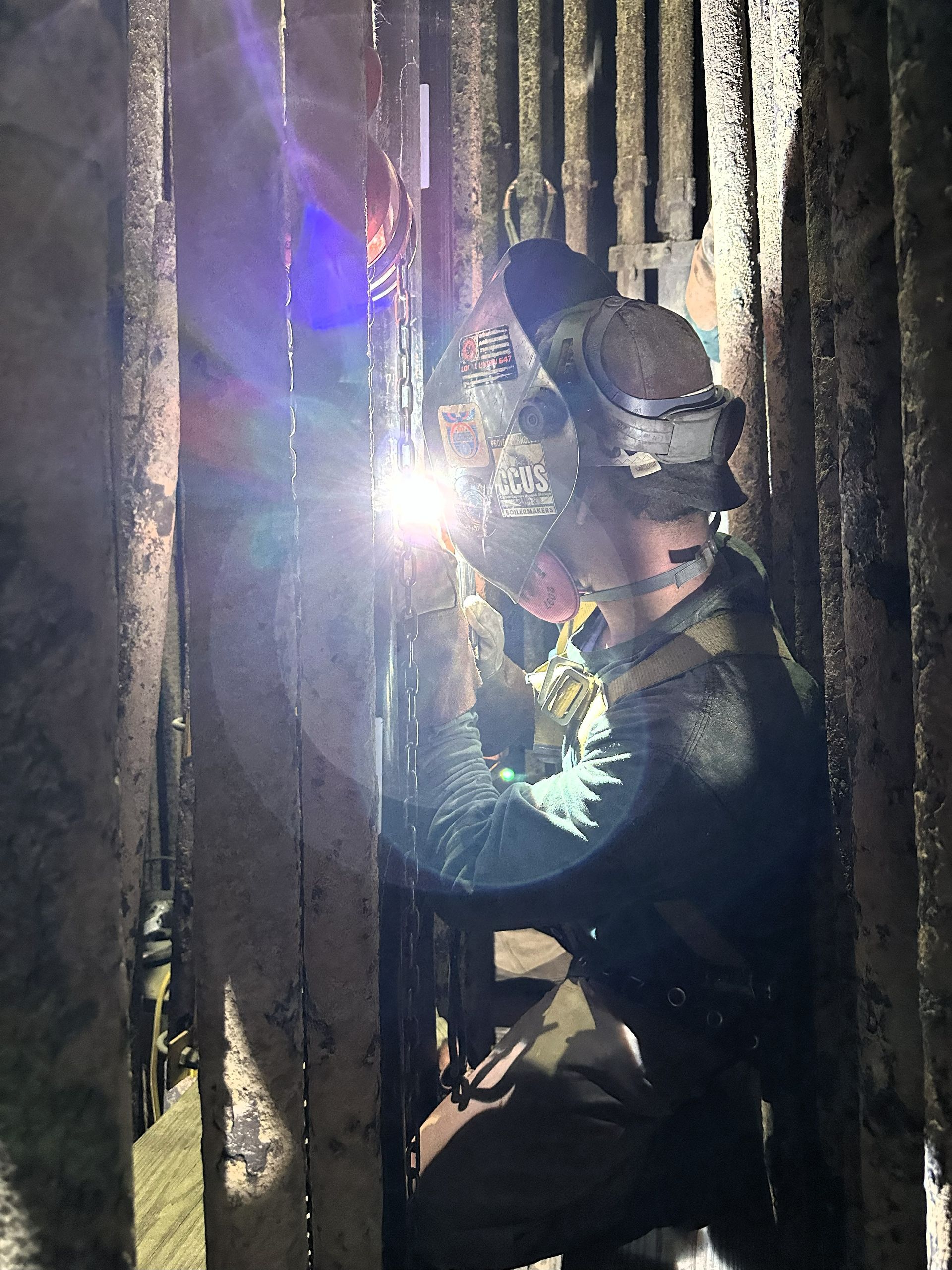 Welder in protective gear welding metal bars in a dimly lit, narrow space. Sparks and bright light are visible.