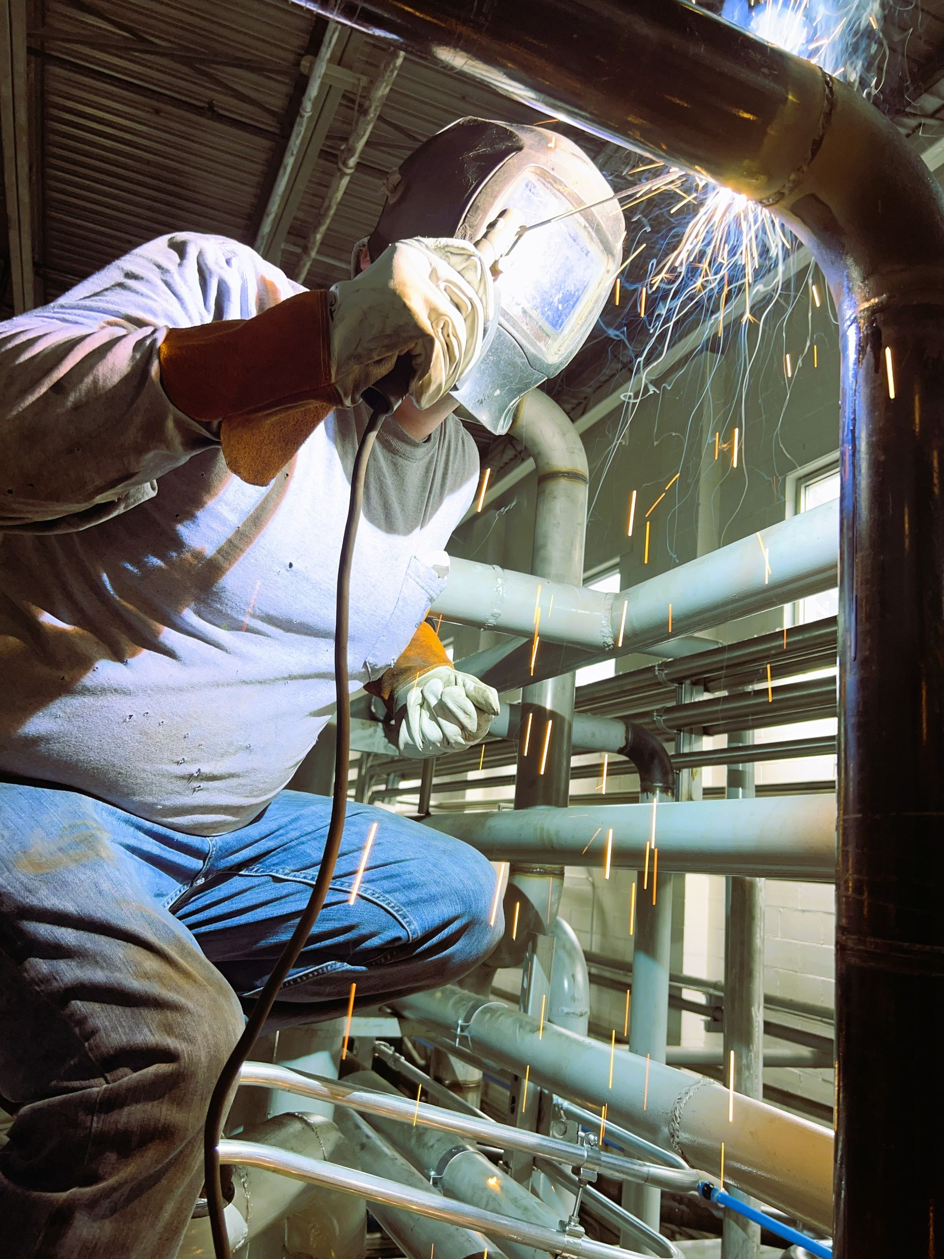 Welder in protective gear welding pipes, sparks flying in an industrial setting.