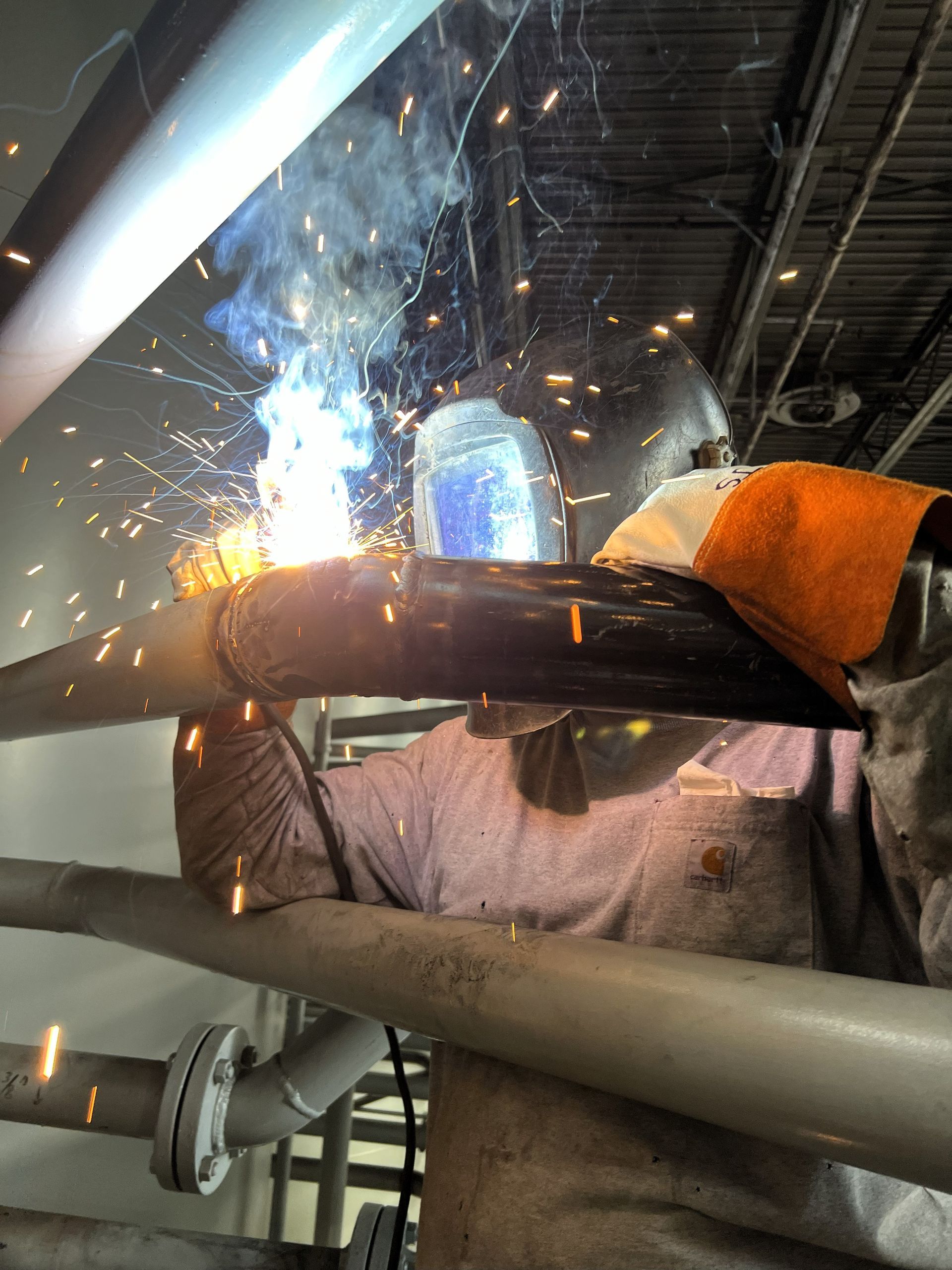 Welder wearing protective gear welding metal pipes, sparks flying in an industrial setting.