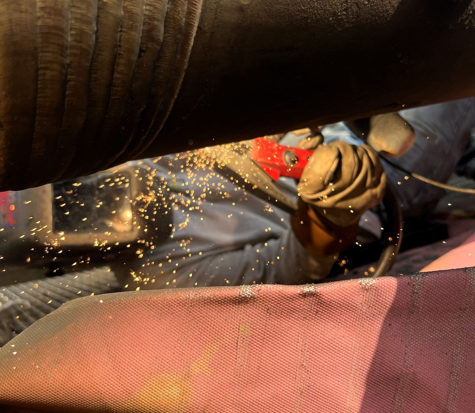 Person welding a pipe, sparks flying. Red-handled valve visible. Protective gear worn.