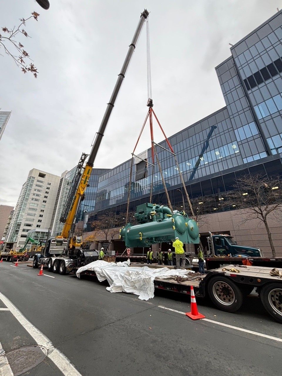 A crane lifts a large green machine over a flatbed truck on a city street. Several workers in yellow vests supervise.