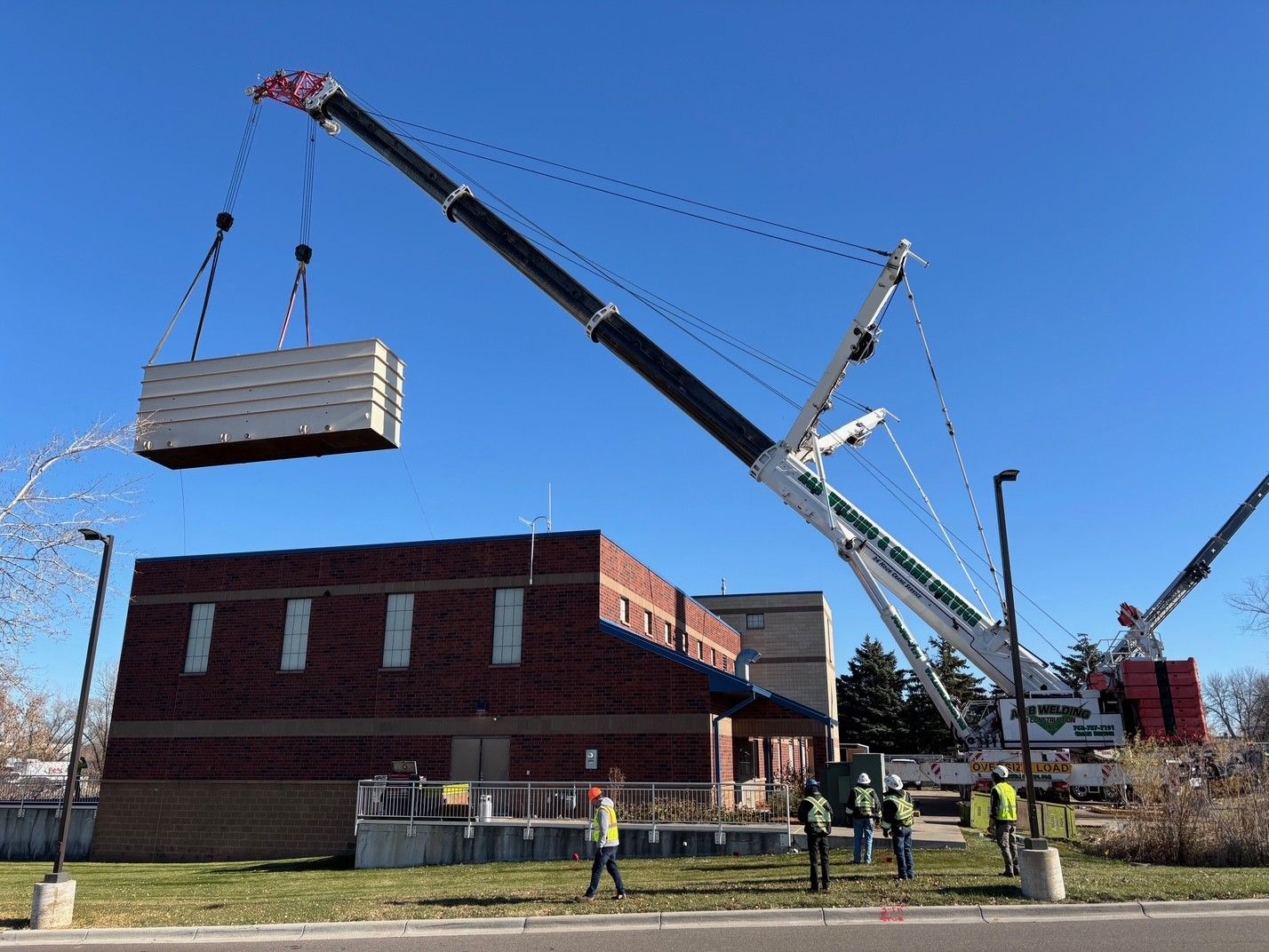 A large crane lifts a rectangular concrete structure above a brick building; several workers in vests look on.
