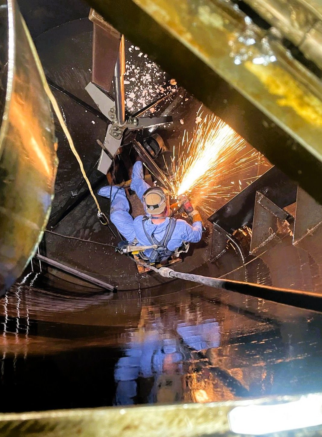Person in blue coveralls welding inside a dark tank, sparks flying.