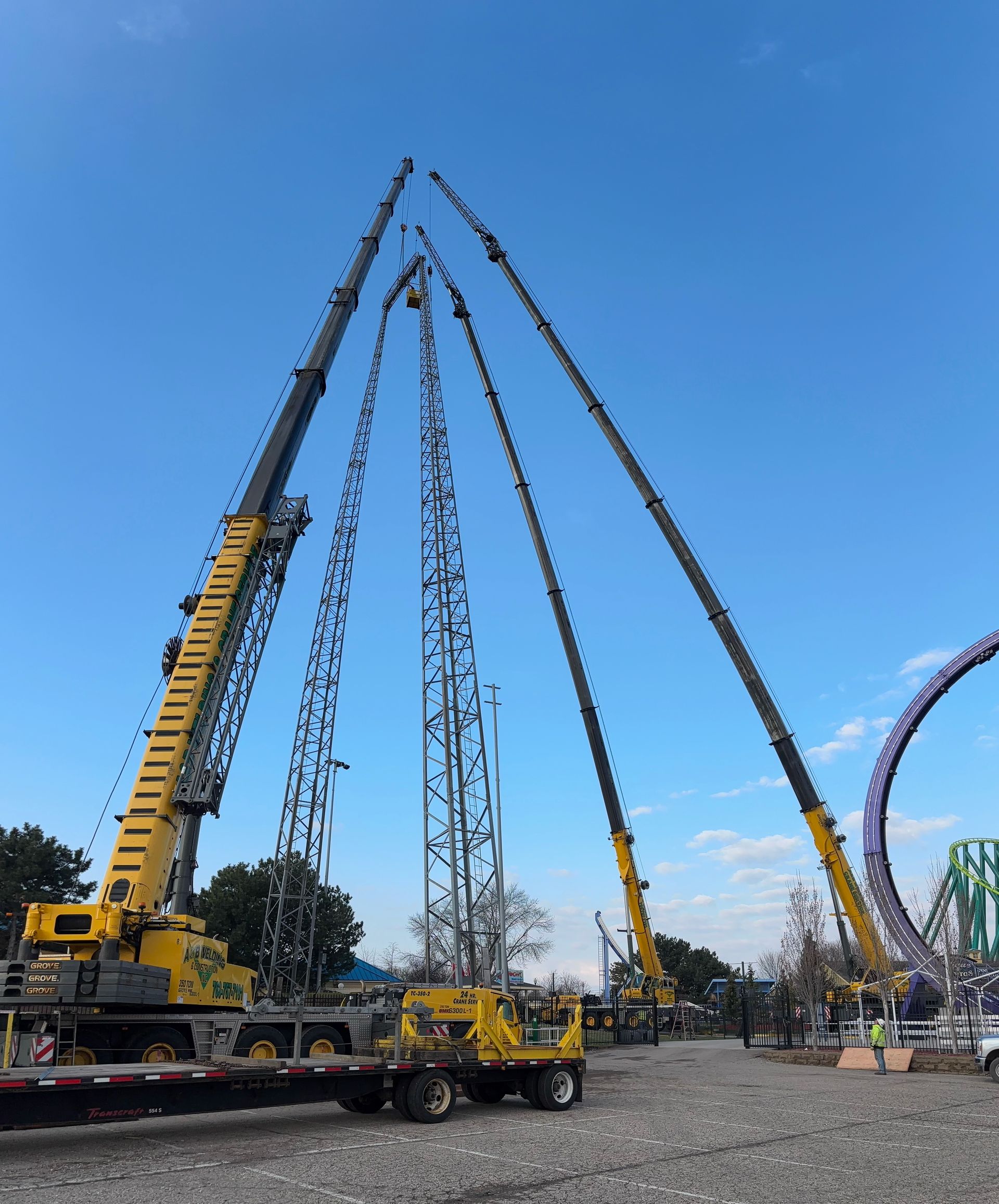 Three large yellow cranes erecting a tall structure, possibly a ride, against a blue sky, near an amusement park.