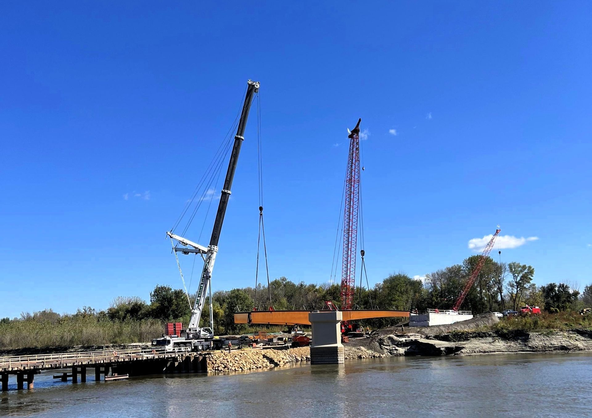Cranes lifting a bridge beam over water during construction on a sunny day.