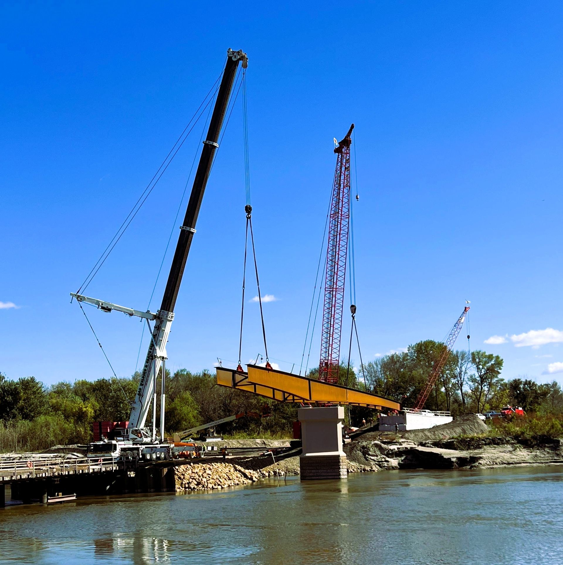 Cranes lifting a bridge beam into place over a river, construction site with blue sky.